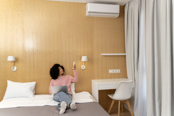 Osoba siedzi na łóżku z laptopem, obsługując klimatyzację do mieszkania za pomocą pilota.